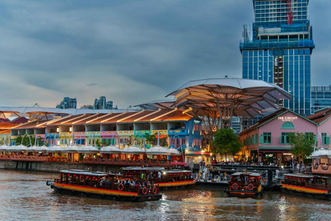 Boats carrying passengers along a river beside pastel-colored waterfront shops and large canopy umbrellas with a tall glass building under construction in the background.