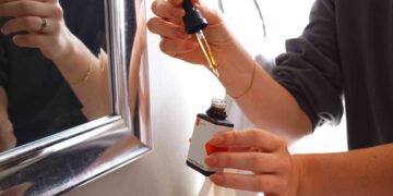 woman-applying-hair-serum-mirror-unsplash - SINwebzine A woman applying anti-frizz serum to damp hair as part of her monsoon beauty routine in a bright bathroom
