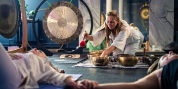 woman-lying-down-singing-bowl-session-unsplash - SINwebzine A woman resting during a sound bath session in a Singapore wellness studio, surrounded by singing bowls and soft light.